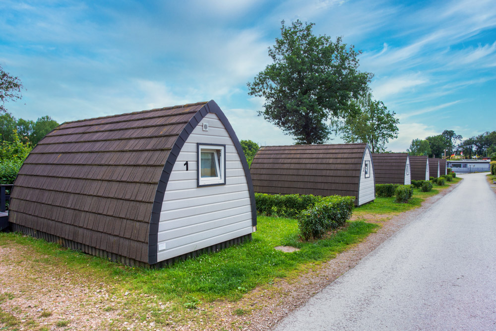 Eine Reihe kleiner Holzhütten mit gewölbten Dächern am Rande einer Schotterstraße, umgeben von Gras und Bäumen, unter einem teilweise bewölkten Himmel.