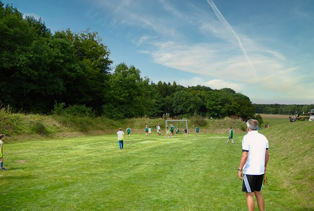 Eine Gruppe von Kindern spielt Fußball auf einer von Bäumen gesäumten Wiese, während ein Erwachsener in Sportkleidung von der Seite zusieht.