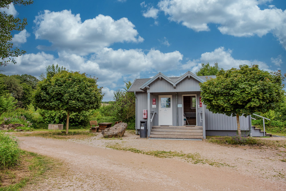 Kleines graues Toilettengebäude mit zwei Eingängen, umgeben von Bäumen und Grünpflanzen, unter einem teilweise bewölkten Himmel auf einem Kiesweg.