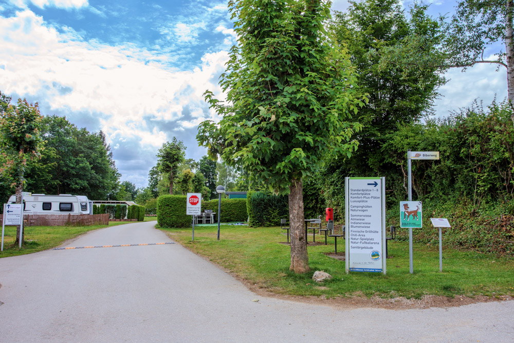 Gepflasterte Straße, die zu einem Campingplatz führt, mit Bäumen, einem geparkten Wohnmobil, Richtungsschildern und einem Stoppschild unter einem teilweise bewölkten Himmel.