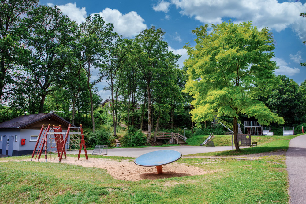 Spielplatz mit einem Klettergerüst, einer runden Drehscheibe in einem Sandkasten, einem Sportplatz, Bäumen und einem kleinen Gebäude unter einem teilweise bewölkten Himmel.