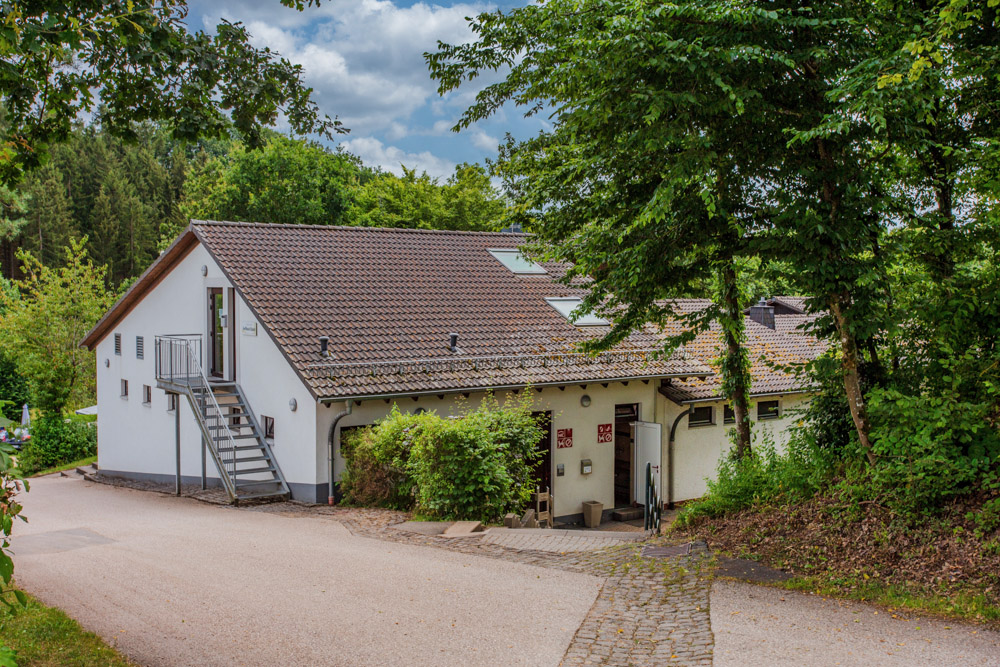 Ein weißes Haus mit schrägem Dach, umgeben von Bäumen und Grünflächen, steht neben einer gepflasterten Straße und einem Kopfsteinpflasterweg unter einem teilweise bewölkten Himmel.