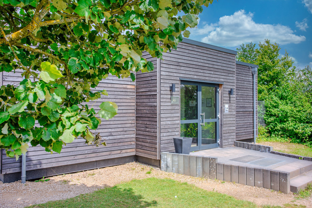 Einstöckiges modernes Gebäude mit Holzverkleidung und einer Glastür, umgeben von Gras, einem Baum und Büschen unter einem blauen Himmel mit Wolken.