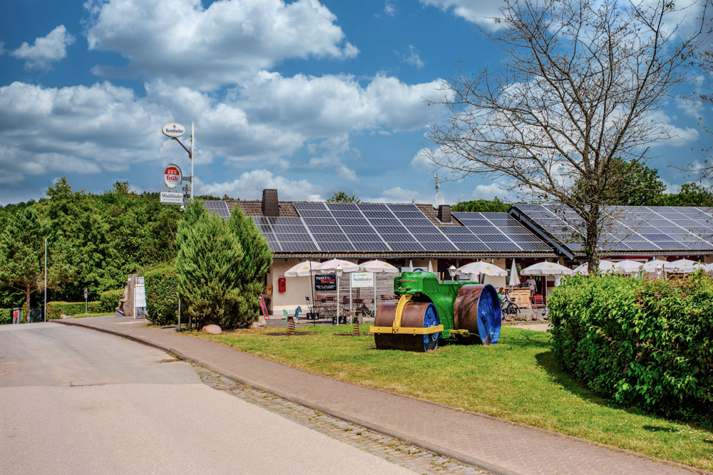 Ein Gebäude mit Sonnenkollektoren auf dem Dach steht in der Nähe einer Straße, mit Sitzgelegenheiten im Freien und zwei bunten rollenden Maschinen auf dem Rasen.