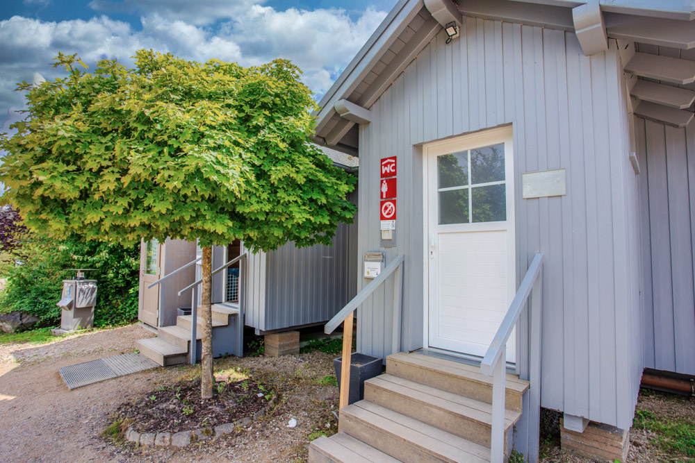 Ein kleines, hellgraues Gebäude mit weißer Tür und Treppe, das neben einem grünen Baum steht, beherbergt Einrichtungen, die durch Schilder am Eingang gekennzeichnet sind.