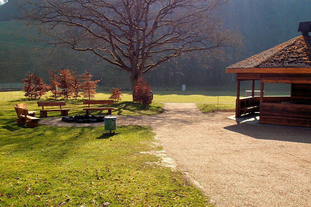 Ein grasbewachsener Parkbereich mit Bänken rund um eine Feuerstelle, einem großen Baum und einem Holzpavillon bei klarem Tageslicht.