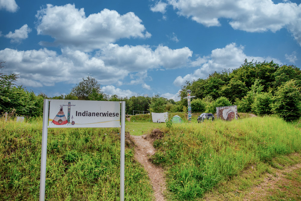 Ein Schild mit der Aufschrift Indianerwiese steht am Eingang zu einer Wiese mit einem Tipi, einem Totempfahl und Bäumen unter einem teilweise bewölkten Himmel.