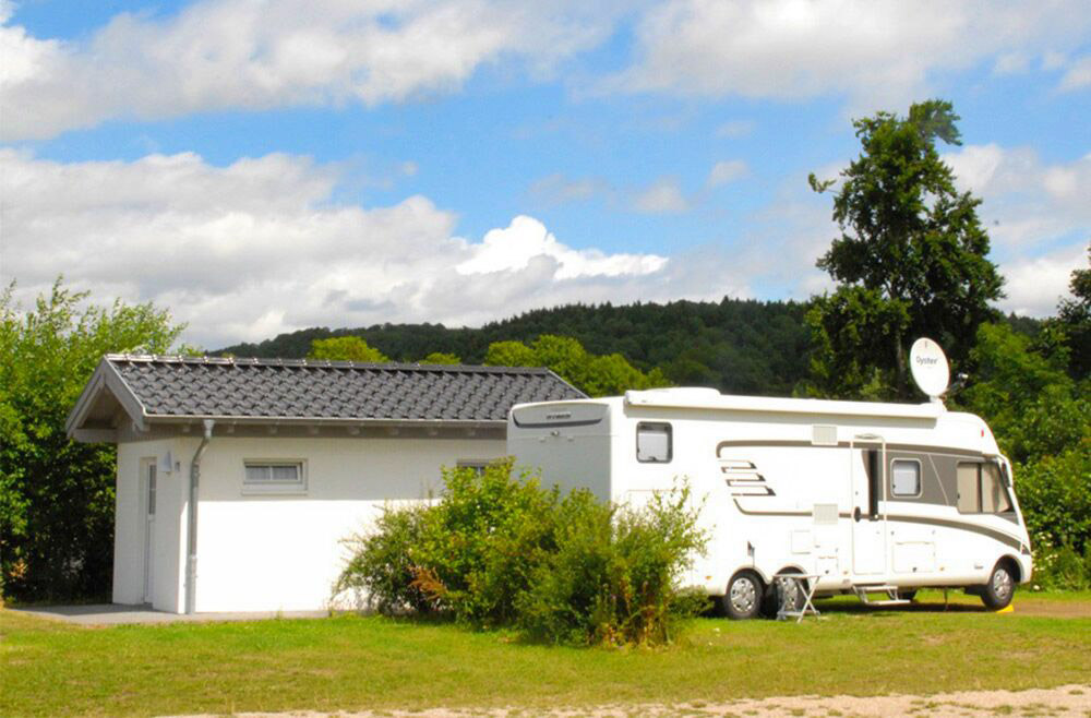 Ein weißes Wohnmobil ist neben einem kleinen weißen Gebäude mit Ziegeldach geparkt, umgeben von grünen Büschen und Bäumen unter einem teilweise bewölkten Himmel.