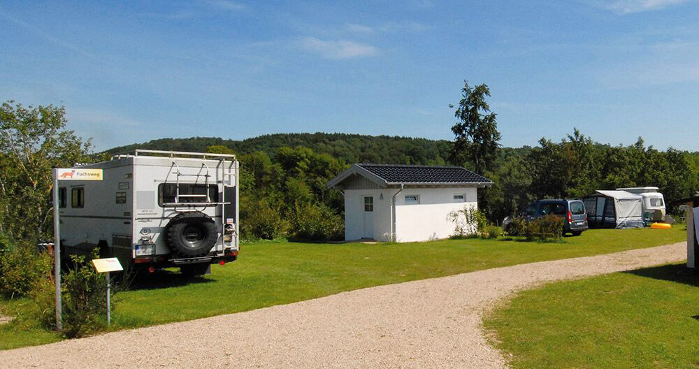 Eine Schotterstraße führt durch einen grasbewachsenen Campingplatz mit einem Wohnmobil, einem kleinen weißen Gebäude und Autos mit Zelten, umgeben von Bäumen unter einem blauen Himmel.