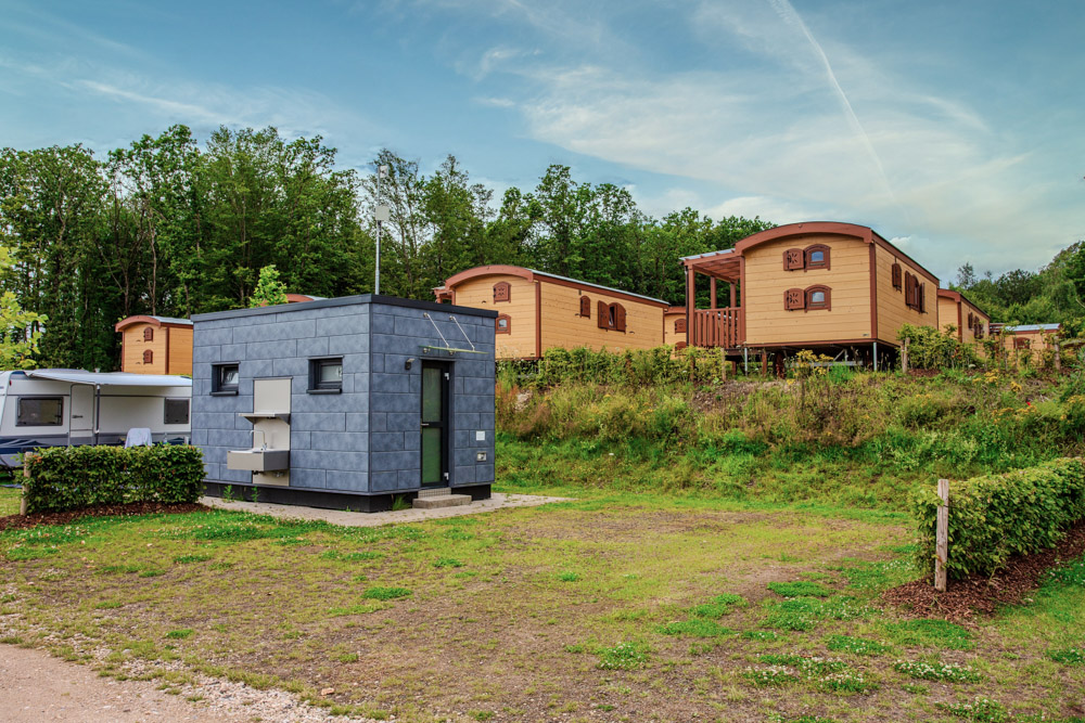 Im Vordergrund befindet sich eine kleine moderne Hütte, im Hintergrund stehen mehrere hölzerne Mobilheime und Bäume unter einem teilweise bewölkten Himmel.