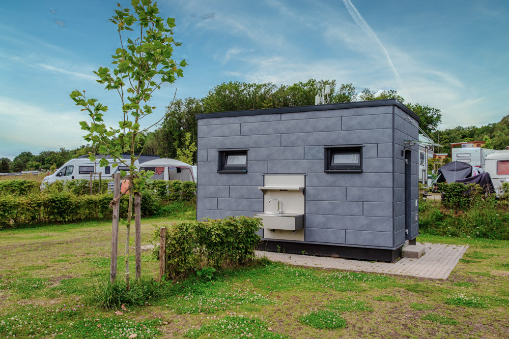 Ein kleines, modernes, graues Gebäude mit zwei Fenstern und einem Außenspülbecken steht auf einem grasbewachsenen Grundstück auf einem Campingplatz mit anderen Wohnmobilen und Bäumen im Hintergrund.