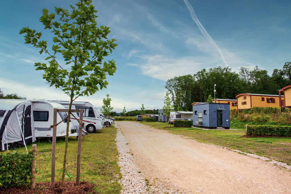 Eine Schotterstraße führt durch einen Campingplatz mit geparkten Wohnwagen, einem kleinen blauen Gebäude, jungen Bäumen und einem strahlend blauen Himmel über uns.