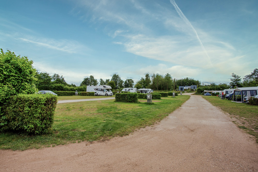 Eine Schotterstraße führt durch einen Campingplatz mit geparkten Wohnmobilen, Zelten und grünen Hecken an einem teilweise bewölkten Tag.