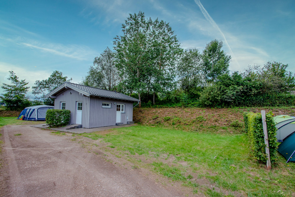 Eine kleine graue Hütte steht auf einem grasbewachsenen Campingplatz mit Zelten in der Nähe, umgeben von Bäumen und unter einem blauen Himmel mit einem sichtbaren Kondensstreifen.