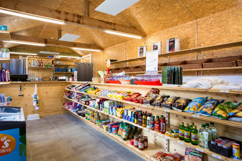 Innenraum eines kleinen Lebensmittelladens mit Holzregalen, die mit Snacks, Konserven, Öl und Getränken gefüllt sind. Der Laden hat eine Holzdecke und einen gefliesten Boden.
