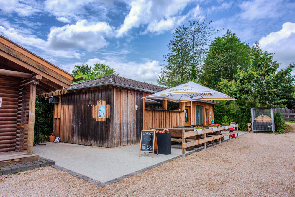 Ein kleines hölzernes Straßencafé mit Menütafeln und einem großen Schirm, umgeben von Bäumen und Kies unter einem teilweise bewölkten Himmel.
