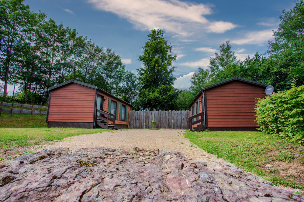 Zwei braune Holzhütten stehen sich gegenüber, dazwischen ein Schotterweg, umgeben von Gras, Bäumen und einem Holzzaun unter einem teilweise bewölkten Himmel.