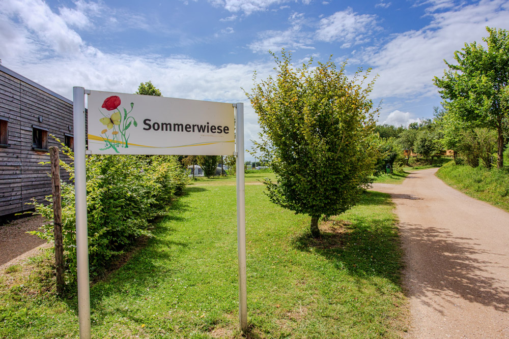 Ein Schild mit der Aufschrift "Sommerwiese" steht auf einer Wiese neben einem unbefestigten Weg, mit Bäumen, Büschen und einem Holzgebäude im Hintergrund unter einem teilweise bewölkten Himmel.
