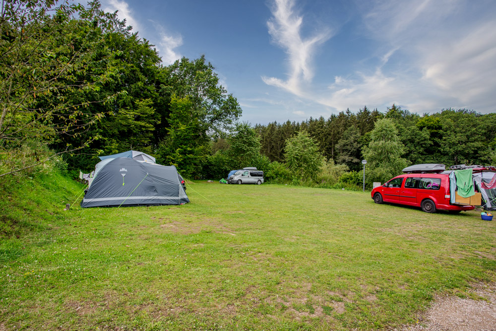 Ein grasbewachsener Campingplatz mit einem aufgestellten Zelt, zwei geparkten Autos und zum Trocknen aufgehängten Kleidungsstücken, umgeben von Bäumen unter einem teilweise bewölkten Himmel.