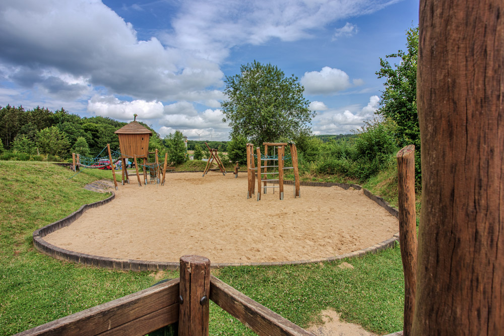 Spielplatz im Freien mit sandigem Untergrund, hölzernen Klettergerüsten und grünem Gras, umgeben von Bäumen und unter einem teilweise bewölkten Himmel.