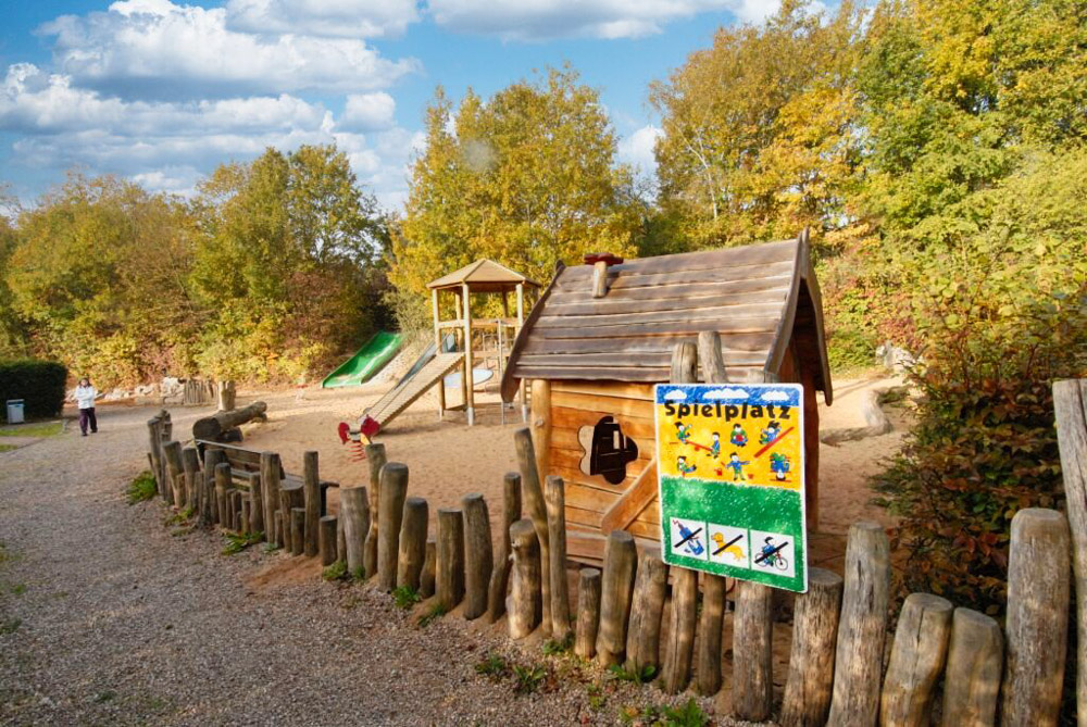 Spielplatz im Freien mit einem Holzspielhaus, einer Rutsche und einem Sandkasten, umgeben von Holzpfosten und Bäumen, mit einem Spielplatzschild im Vordergrund.
