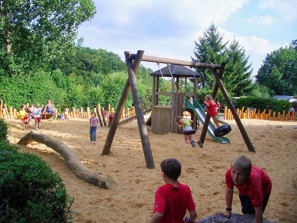 Kinder spielen auf Schaukeln und Rutschen auf einem Sandspielplatz, der von Bäumen und einem Holzzaun umgeben ist, während die Erwachsenen im Hintergrund sitzen.