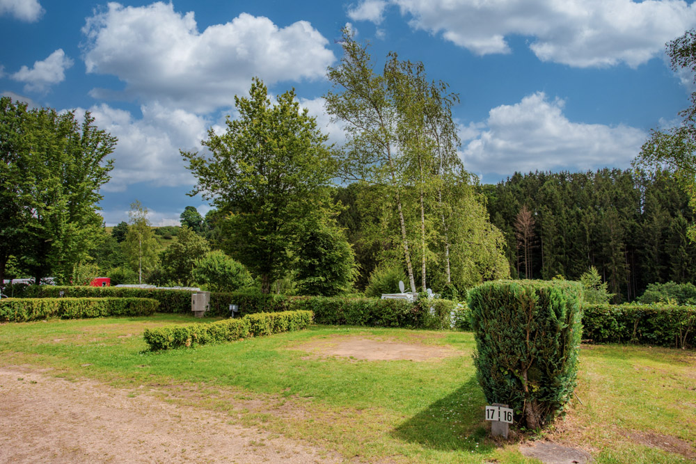 Ein grasbewachsener Campingplatz mit beschnittenen Büschen, umgeben von Bäumen und Wald unter einem teilweise bewölkten Himmel. Ein nummeriertes Schild mit der Aufschrift 17 | 16 steht in der Nähe der Hecke.