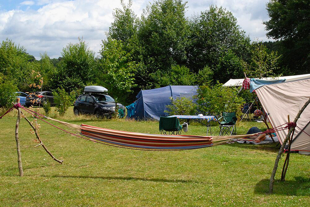 Eine gestreifte Hängematte hängt zwischen zwei Holzpfosten im Gras, im Hintergrund stehen Zelte, Campingstühle und Autos, umgeben von grünen Bäumen.