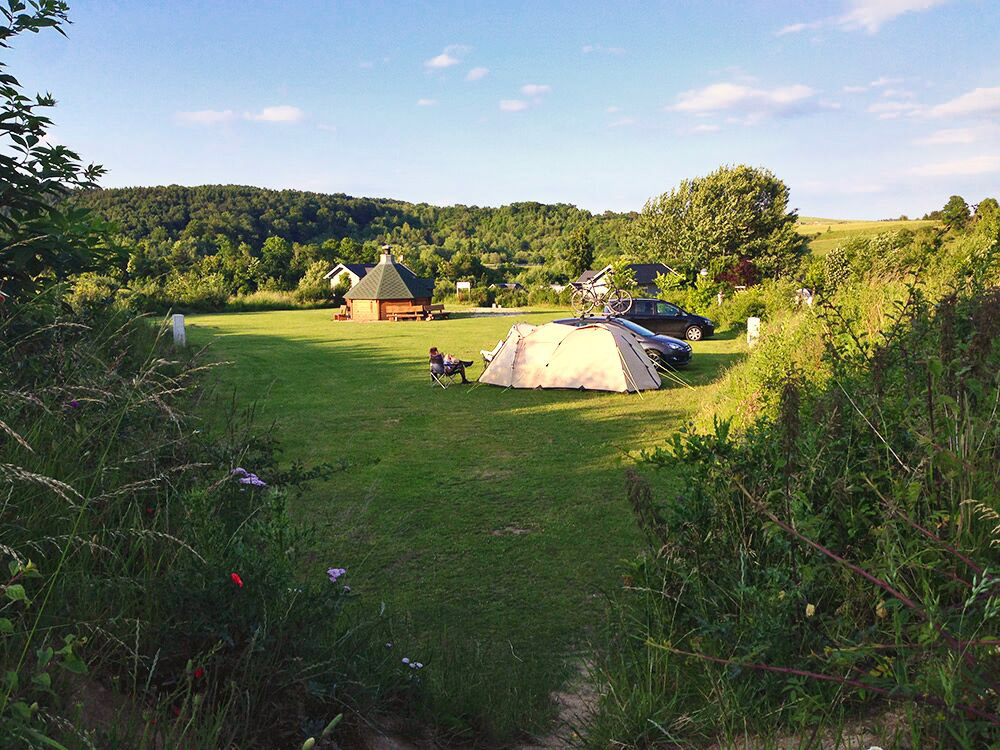 Ein Campingplatz auf einer Wiese mit einem Zelt, geparkten Autos, einer kleinen Holzhütte und einer auf einem Stuhl sitzenden Person; im Hintergrund sind Bäume und Hügel unter einem blauen Himmel zu sehen.