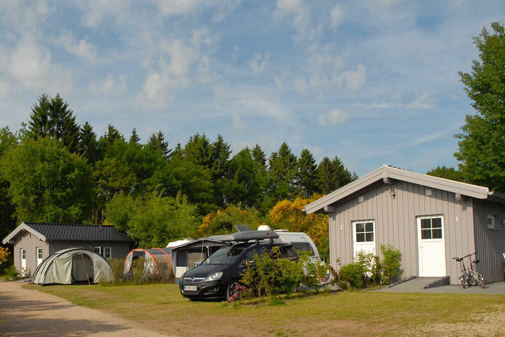 Eine kleine graue Hütte, ein Auto, Zelte und ein Wohnwagen sind auf einer Wiese in der Nähe eines Waldgebiets unter einem teilweise bewölkten Himmel aufgebaut.