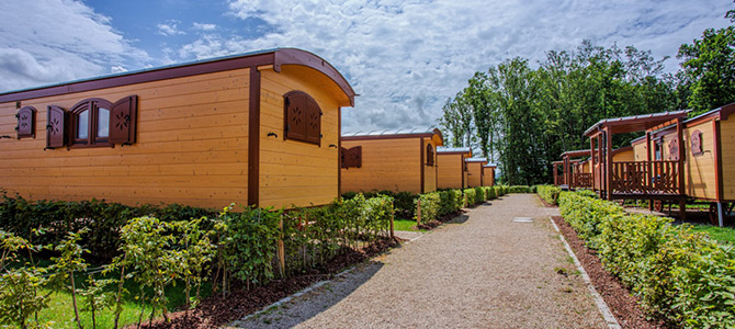 Verschillende houten hutten met decoratieve luiken staan op een rij langs een grindpad, omringd door groene struiken en bomen onder een gedeeltelijk bewolkte hemel.