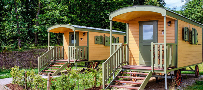 Twee kleine houten hutten op wielen met groene deuren en luiken, elk met een trapje dat leidt naar een veranda, omringd door groen en bomen.