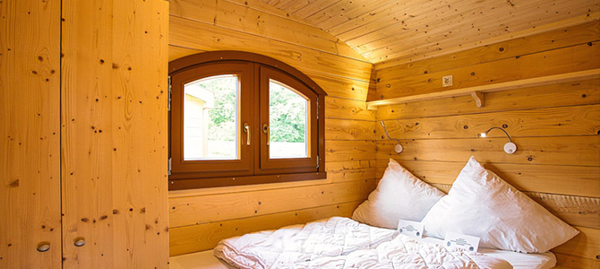 Kleine houten slaapkamer met een tweepersoonsbed, wit beddengoed, een boograam, ingebouwde kledingkast, muurverlichting en een plank boven het bed.