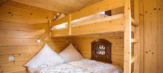 Een kleine houten kamer met een stapelbed; het onderste bed is een tweepersoonsbed met wit beddengoed en het bovenste bed is een eenpersoonsbed. Er is een klein raam en leeslampjes aan de muur.