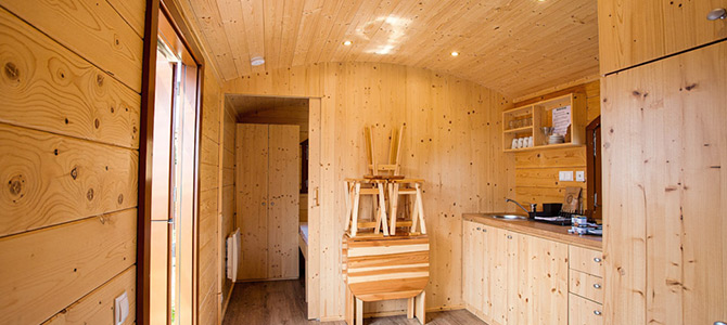 Kleine houten hut met licht grenen muren, gestapelde stoelen op een klaptafel, een compact keukentje, planken met serviesgoed en een gang die naar een andere kamer leidt.