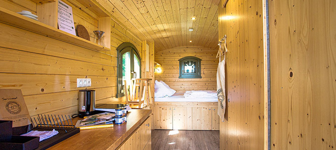 Interieur van een kleine houten hut met planken, een aanrecht, twee ramen en een bed aan het uiteinde; overal natuurlijke houtafwerking.