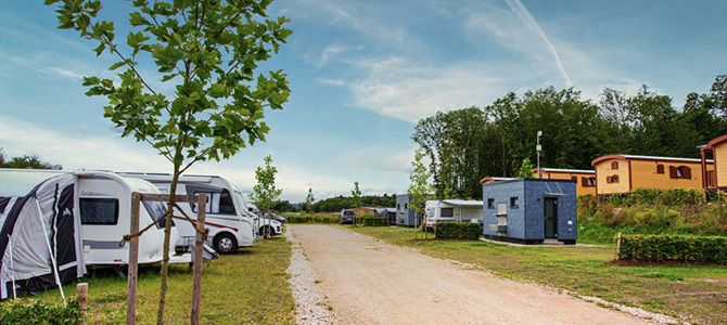 Eine Schotterstraße führt durch einen Campingplatz mit geparkten Wohnwagen, kleinen Wirtschaftsgebäuden, grünem Gras, jungen Bäumen und Hütten im Hintergrund unter einem teilweise bewölkten Himmel.
