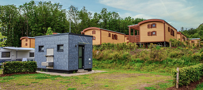 Im Vordergrund steht ein kleines, modernes, graues Häuschen, im Hintergrund stehen mehrere Holzhütten und Bäume auf einem grasbewachsenen Hügel.
