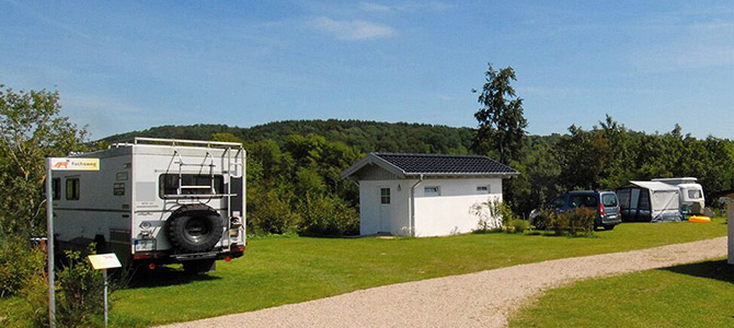 Ein kleiner Campingplatz mit geparkten Wohnmobilen, einem weißen Versorgungsgebäude, Grasflächen und Bäumen im Hintergrund unter einem klaren Himmel.