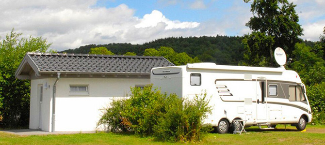 Ein weißes Wohnmobil ist neben einem kleinen weißen Gebäude auf einer Wiese geparkt, mit Bäumen und einem teilweise bewölkten Himmel im Hintergrund.