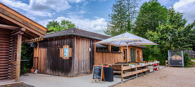 Ein kleines Holzgebäude mit einer überdachten Terrasse, Sitzgelegenheiten im Freien, einem großen Sonnenschirm und einem Kreideschild, umgeben von Bäumen und einem teilweise bewölkten Himmel.