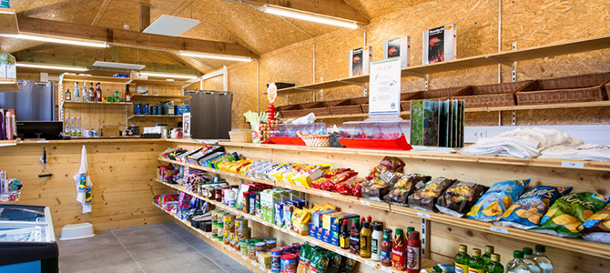 Kleiner Lebensmittelladen mit Holzregalen mit Snacks, Konserven, Getränken und anderen Lebensmitteln. Im Hintergrund sind Theke und Kühlregal zu sehen.