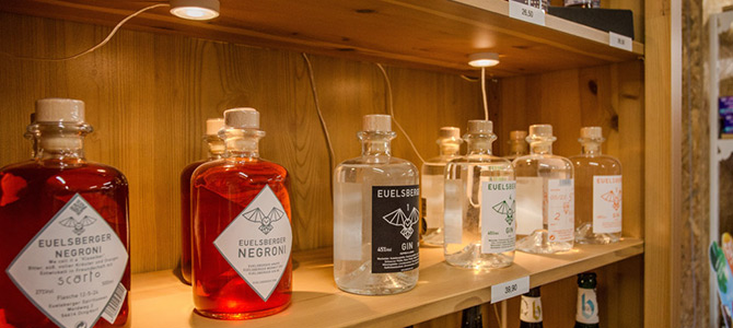 Die Flaschen verschiedener Euelsberger Negroni-Spirituosen stehen auf Holzregalen, über denen kleine Lichter leuchten.