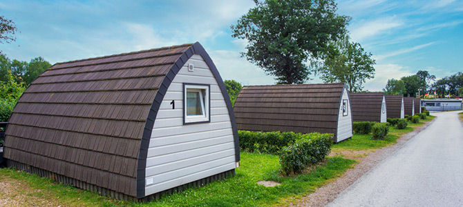Eine Reihe von kleinen, identischen Holzhütten mit braunen Dächern und weißen Wänden entlang eines Kiesweges, umgeben von Grün und Bäumen unter blauem Himmel.