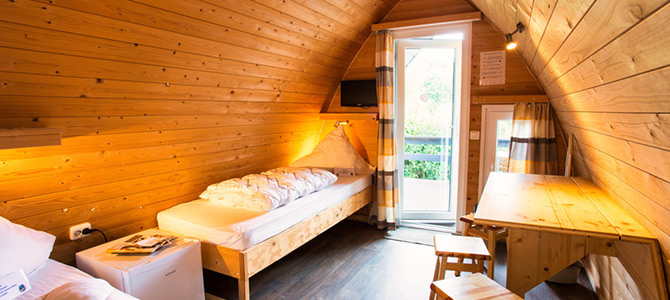 Eine kleine Holzhütte mit schrägen Wänden, einem Einzelbett, einem Tisch mit Hockern, einem Minikühlschrank, einem an der Wand befestigten Fernseher und einer offenen Tür nach draußen.