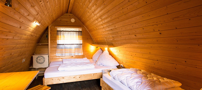 Innenraum einer kleinen Holzhütte mit schräger Decke, zwei ungemachten Betten, einem Tisch, einem Fenster mit Vorhängen und warmer Beleuchtung.