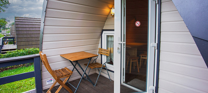 Kleine Holzterrasse mit einem Tisch und zwei Stühlen vor einer modernen Hütte; Glastür führt ins Innere, Rauchverbotsschild sichtbar, Grün im Hintergrund.