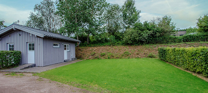 A small gray house with a white door and windows sits beside a neatly trimmed lawn, bordered by hedges and trees under a cloudy sky.