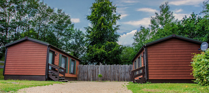 Two brown wooden cabins, one featuring a cozy sauna, face each other across a gravel path, with a wooden fence and tall trees in the background under a partly cloudy sky.
