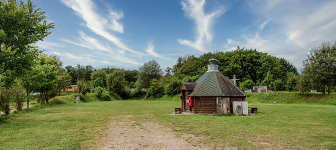 Eine kleine Holzhütte mit einem sechseckigen Dach steht inmitten eines grasbewachsenen Feldes, umgeben von Bäumen unter einem teilweise bewölkten blauen Himmel.